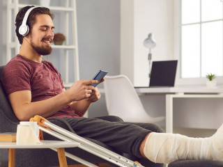 Young man with broken leg sitting in chair and listening to music on mobile phone