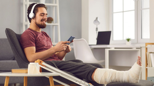 Young man with broken leg sitting in chair and listening to music on mobile phone