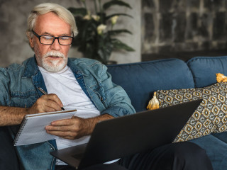 Focused elderly man writing notes in notebook watching webinar or online training using laptop computer modern senior male with gray hair and beard learning online