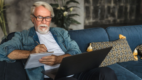 Focused elderly man writing notes in notebook watching webinar or online training using laptop computer modern senior male with gray hair and beard learning online