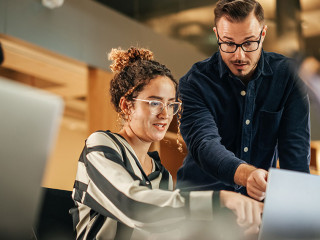 Two Colleagues Working On Laptop in Office. Female Hispanic Help