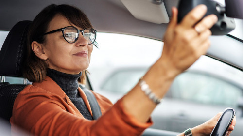 Adult caucasian businesswoman in glasses adjusting the rearview mirror while sitting at the wheel of a car