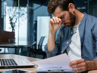 Upset businessman behind paper work inside modern office, mature man with beard reading financial reports and account documents unhappy with results and disappointed with achievements