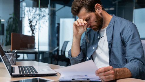 Upset businessman behind paper work inside modern office, mature man with beard reading financial reports and account documents unhappy with results and disappointed with achievements
