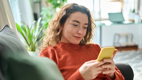 Smiling relaxed young woman sitting on couch using mobile phone technology.