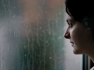Woman standing by window during rainy day looking outside watchi