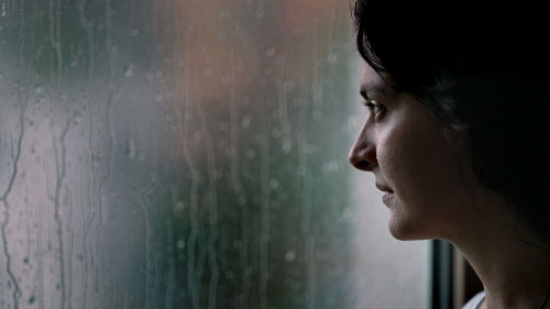 Woman standing by window during rainy day looking outside watchi