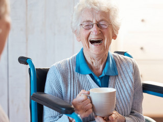 Senior woman, friend and coffee or tea while in a wheelchair for a disability or rehabilitation and happy or laughing about funny conversation or joke. Old people together for fun in nursing home