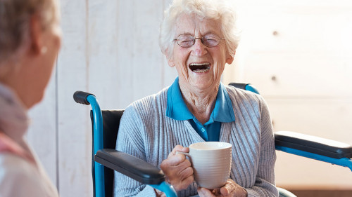 Senior woman, friend and coffee or tea while in a wheelchair for a disability or rehabilitation and happy or laughing about funny conversation or joke. Old people together for fun in nursing home