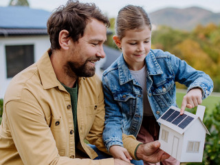 Little girl with her dad holding paper model of house with solar panels, explaining how it works.Alternative energy, saving resources and sustainable lifestyle concept.