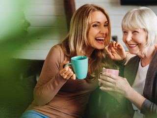 Happy senior mother with adult daughter sitting on couch and holding cups with coffee or tea at home. Togetherness concept