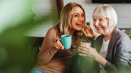 Happy senior mother with adult daughter sitting on couch and holding cups with coffee or tea at home. Togetherness concept