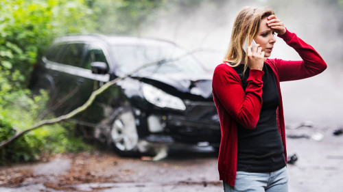 A young woman with smartphone by the damaged car after a car accident, making a phone call.