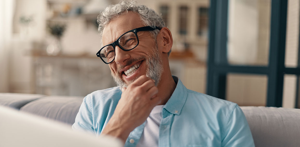 Cheerful senior man in casual clothing and eyeglasses using lapt
