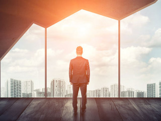 Businessman standing on the floor back to us looking through the big window with cityscape and sunset behind it.