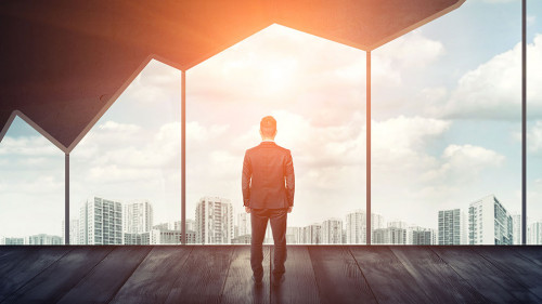 Businessman standing on the floor back to us looking through the big window with cityscape and sunset behind it.