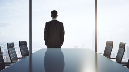 Empty meeting room with big conference table, chairs around and businessman back looking through big window on bright sky.