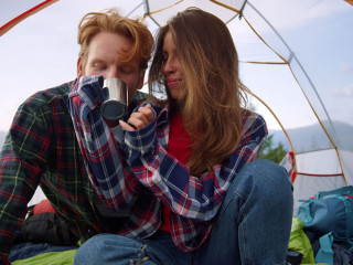 Young couple sitting in tent during hike. Redhead guy drinking tea from cup