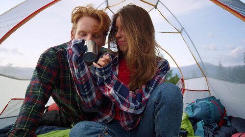Young couple sitting in tent during hike. Redhead guy drinking tea from cup
