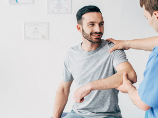 Smiling patient sitting on couch and doctor examining patient shoulder in massage cabinet at clinic