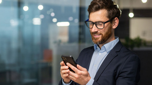 Successful financier investor works inside office at work, businessman in business suit uses telephone near window, man smiles and reads good news online from smartphone
