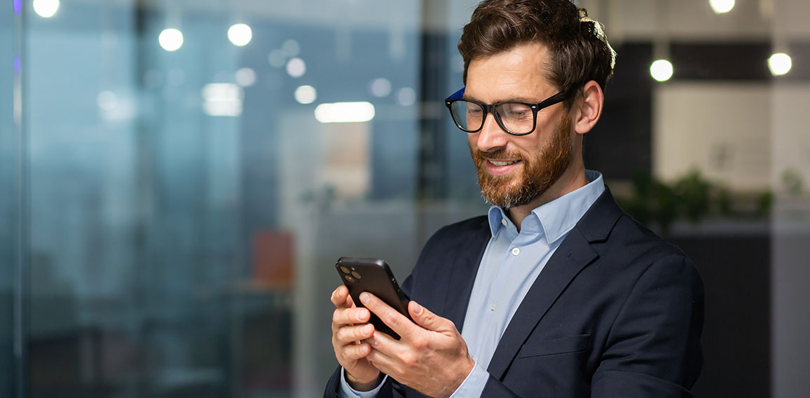 Successful financier investor works inside office at work, businessman in business suit uses telephone near window, man smiles and reads good news online from smartphone