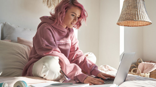 Hipster teen girl student with pink hair using laptop online learning in bed.