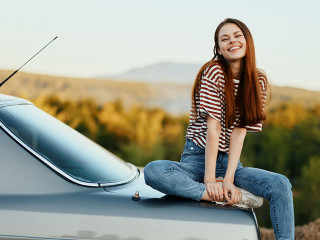 A woman car driver sits on the trunk of a car and smiles admiring a beautiful view of autumn nature and mountains