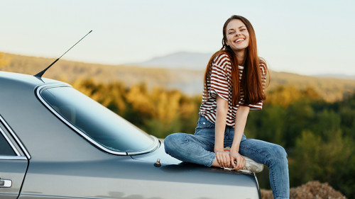 A woman car driver sits on the trunk of a car and smiles admiring a beautiful view of autumn nature and mountains