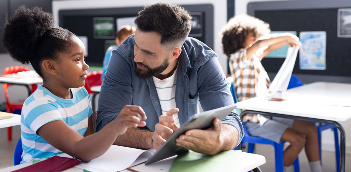 Diverse male teacher and schoolgirl using tablet at her desk in