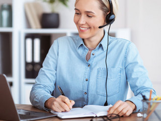 Happy young woman in headset participating in online lesson or web conference, taking notes, studying remotely
