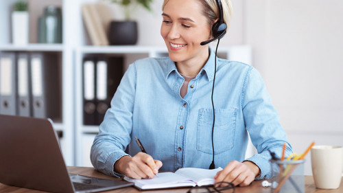 Happy young woman in headset participating in online lesson or web conference, taking notes, studying remotely