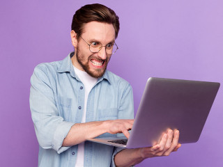 Photo of unhappy angry young man look hold laptop write letter work isolated on purple color background