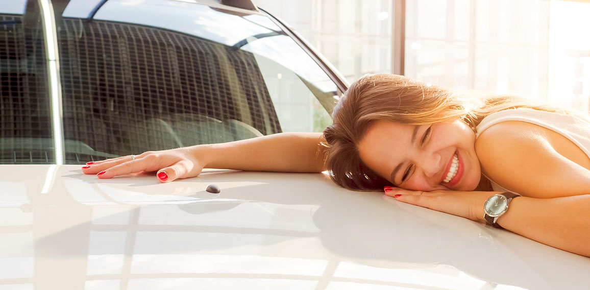 Dream about car. Gorgeous smiling woman hugging lies on the hood of new white car in the dealership.