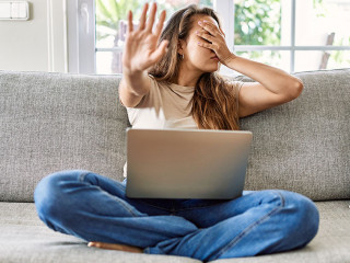 Beautiful young brunette woman sitting on the sofa using computer laptop at home covering eyes with hands and doing stop gesture with sad and fear expression. embarrassed and negative concept.