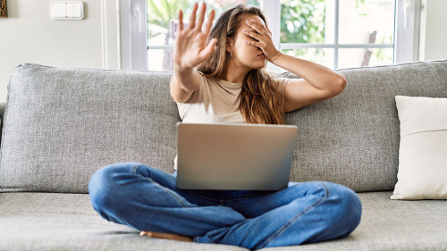 Beautiful young brunette woman sitting on the sofa using computer laptop at home covering eyes with hands and doing stop gesture with sad and fear expression. embarrassed and negative concept.