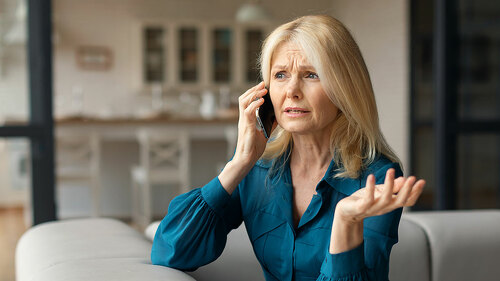 Stressed mature woman talking on cellphone, having unpleasant phone conversation while sitting on sofa at home