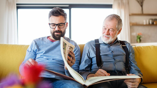 An adult hipster son and senior father sitting on sofa indoors at home.
