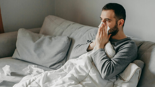 Young sick bearded european man sneezes into napkin at home on g