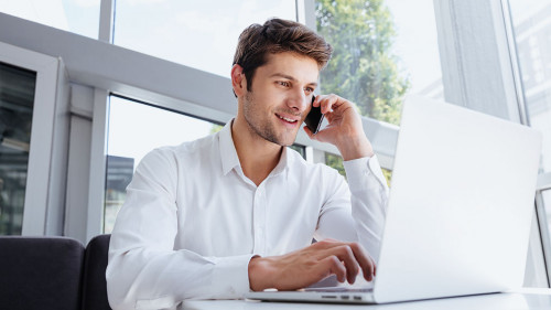 Happy young businessman talking on cell phone and using laptop