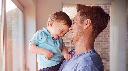 Father holds toddler son while drinking coffee at home, by the w