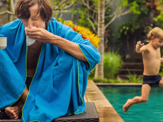 Sick man traveler. The man caught a cold on vacation, sits sad at the pool drinking tea and blows his nose into a napkin. His son is healthy and swimming in the pool. Travel insurance concept