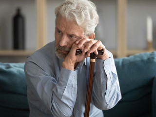 Tired upset mature man holding walking stick, sitting on couch