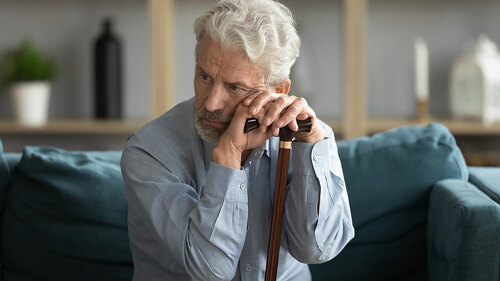 Tired upset mature man holding walking stick, sitting on couch