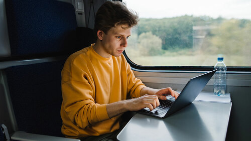 Young male freelancer in orange sweatshirt working on laptop in