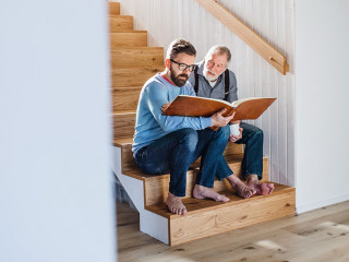 An adult son and senior father sitting on stairs indoors at home, looking at photographs.