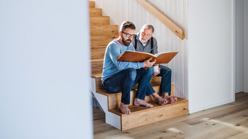 An adult son and senior father sitting on stairs indoors at home, looking at photographs.