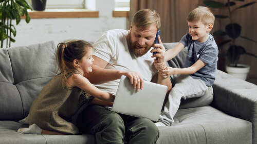 Man, father sitting on couch, talking on phone and working on laptop in living room. Kids bothering and playing with him