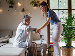 Side view of healthcare worker or caregiver visiting senior woman indoors at home, helping her to walk.