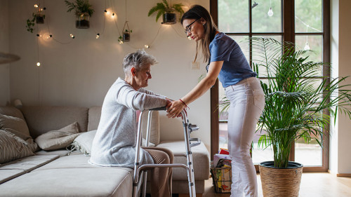 Side view of healthcare worker or caregiver visiting senior woman indoors at home, helping her to walk.